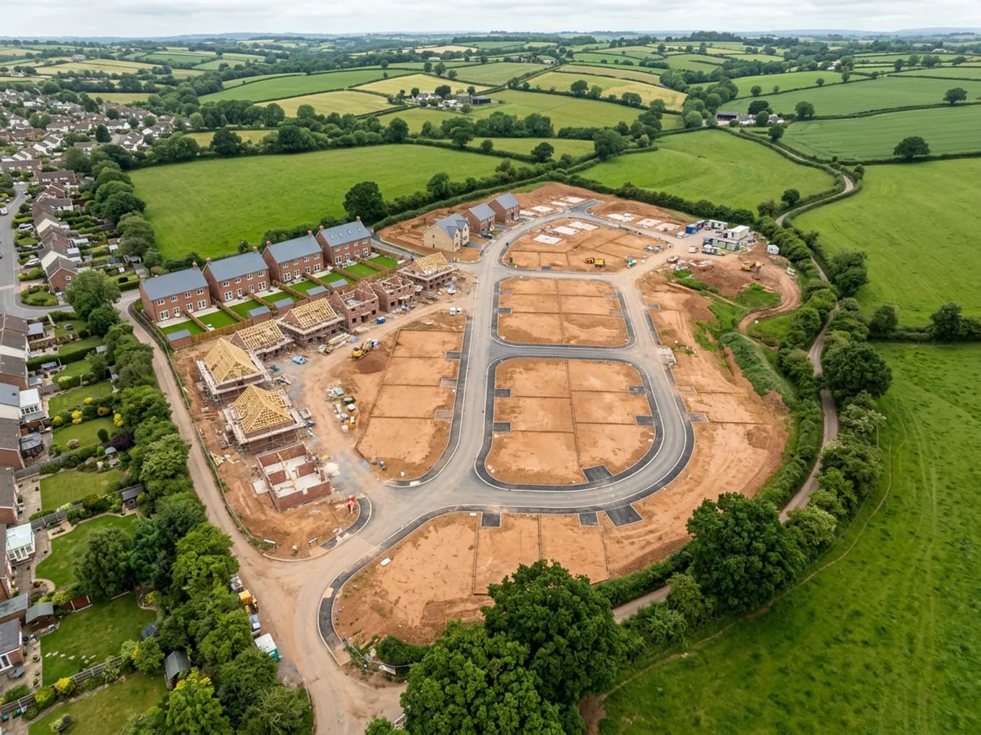 A straight down aerial view of a part built housing development in Devon, showing plot outlines, new road layout, stockpiled topsoil and the surrounding farmland boundaries