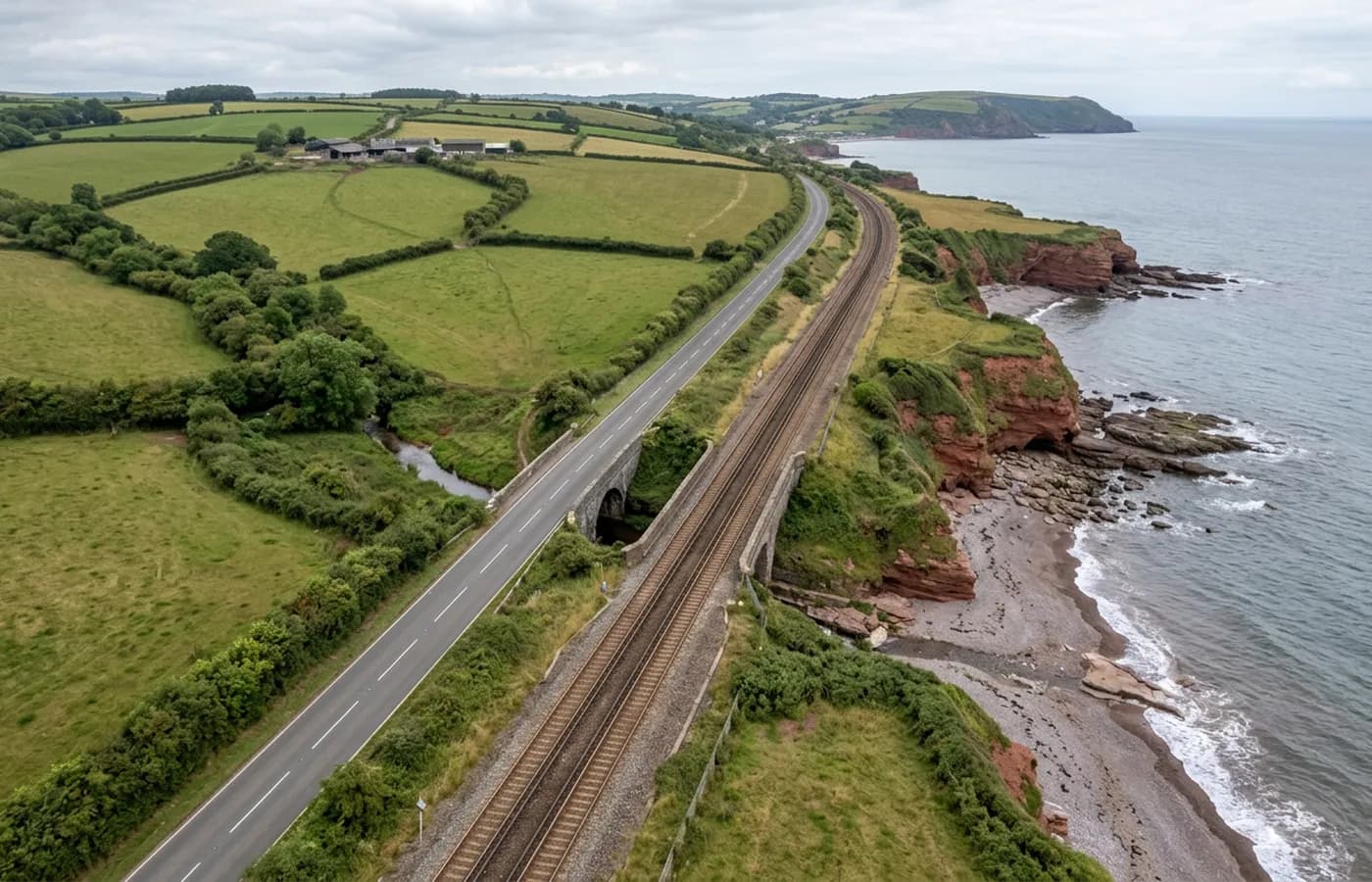 An aerial view along a transport corridor in Devon, road and railway running side by side between the coast and farmland